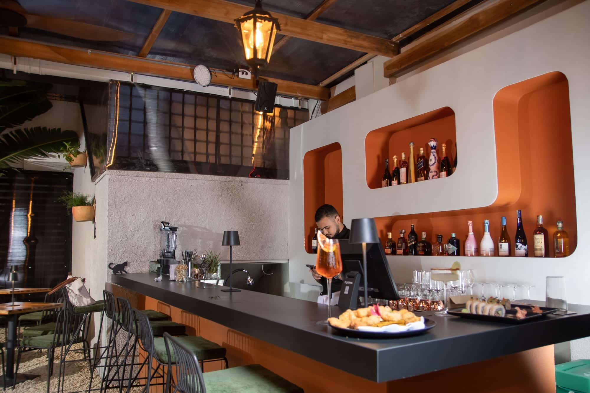 The patio bar: warm-lit niches with bottles, the bartender and a freshly poured spritz