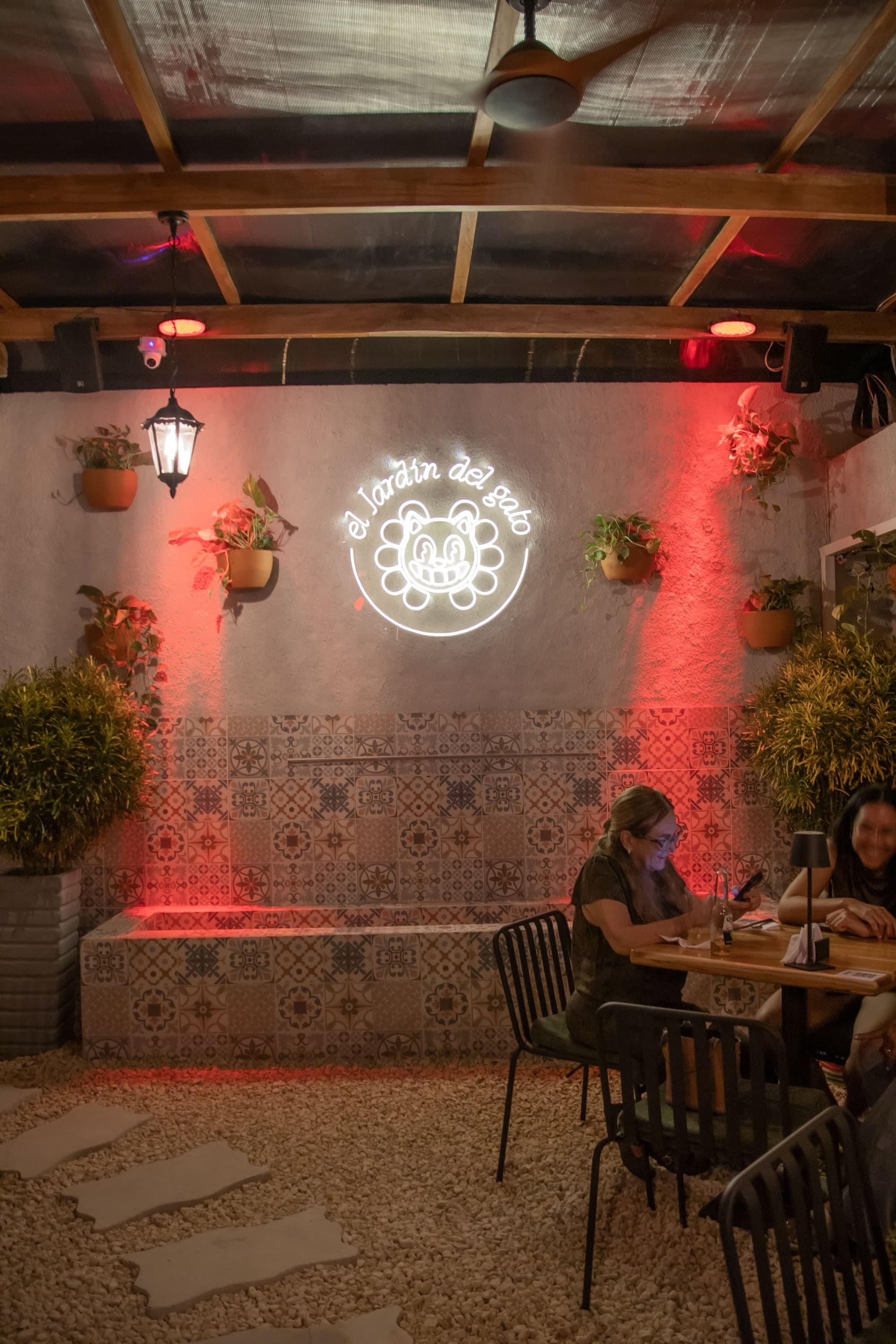 The patio at El Jardín del Gato at night: tables on the gravel, hanging pots, lanterns and the neon cat sign at the back