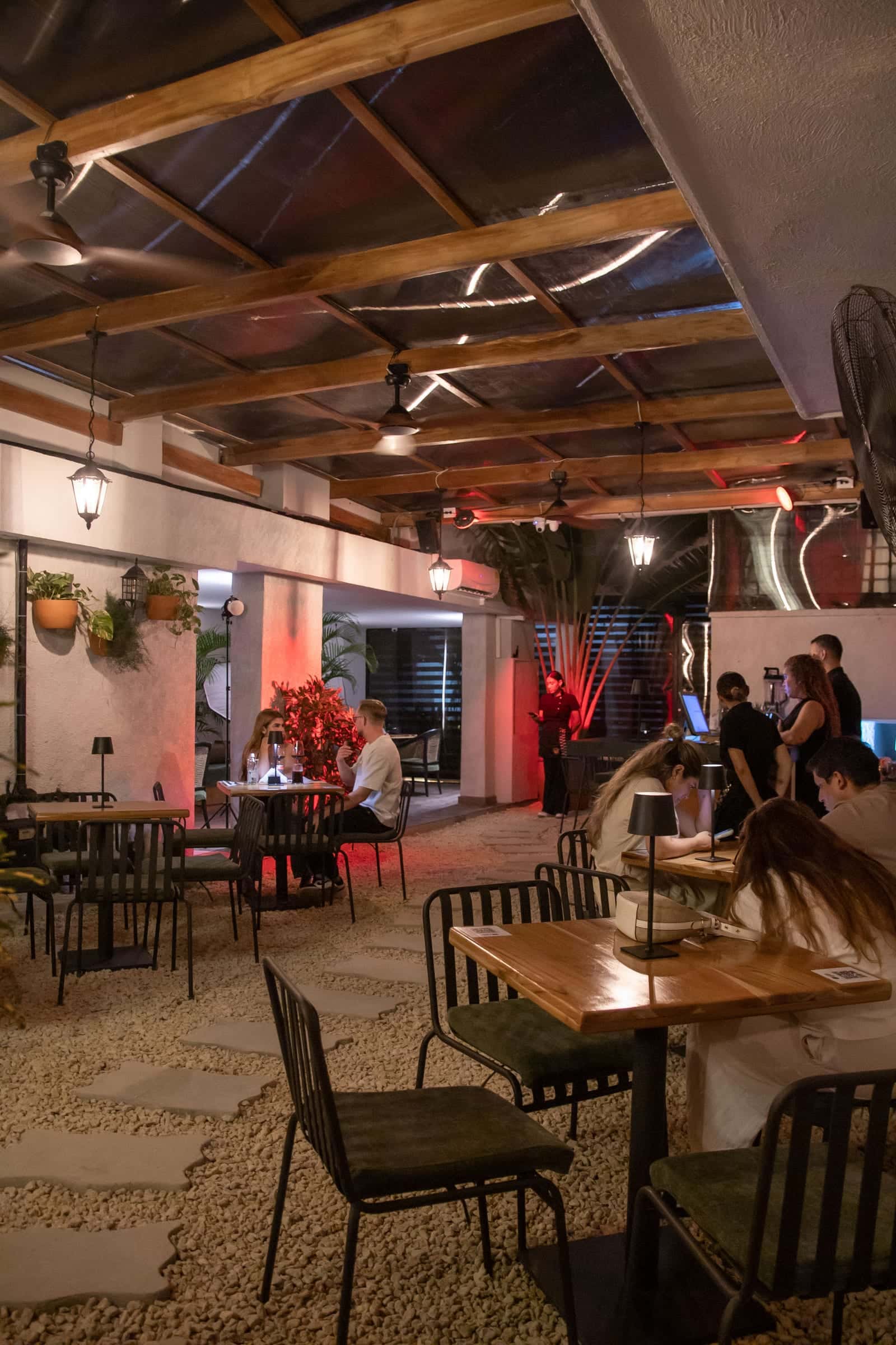 The patio at night in depth: tables on the gravel, lanterns, plants and guests at the back