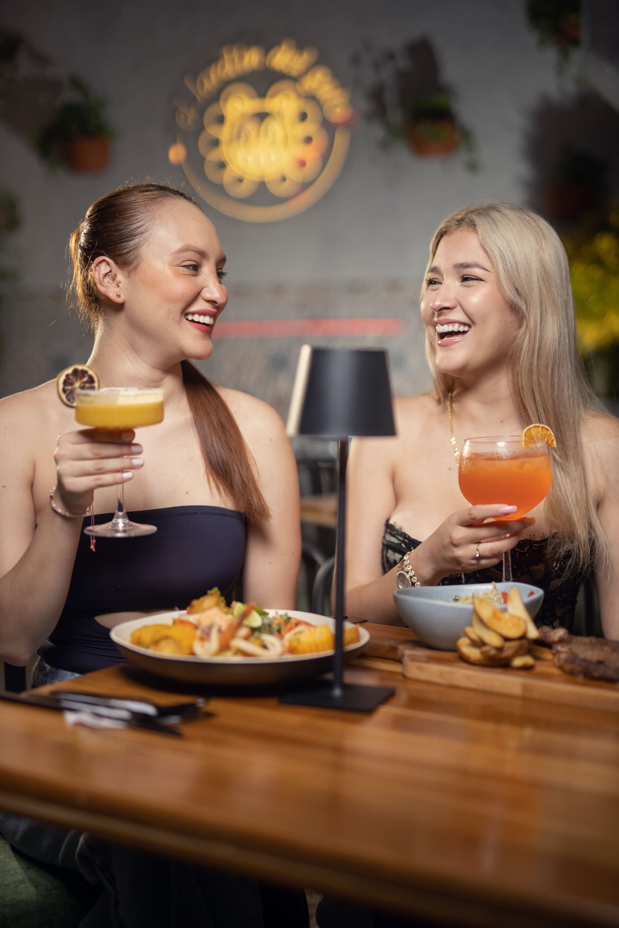 Two friends laugh over their cocktails and dishes at a table, under the cat logo